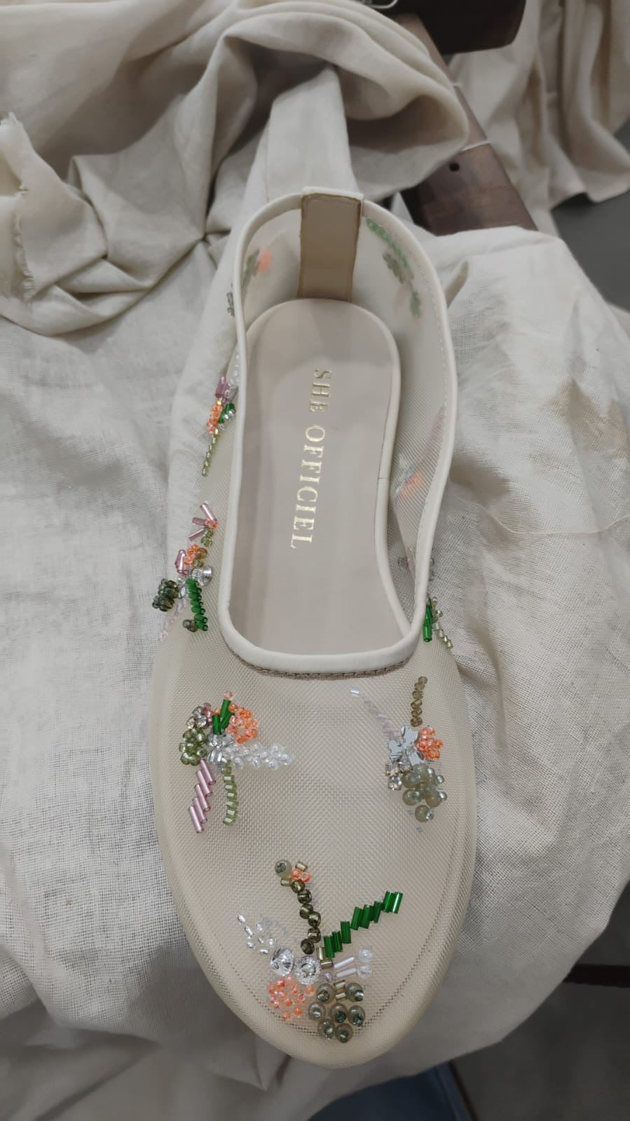 A single cream-colored slip-on shoe with floral embroidery and beadwork, resting on a draped beige fabric background. The insole displays the brand name 'SHI OFFICIAL'.