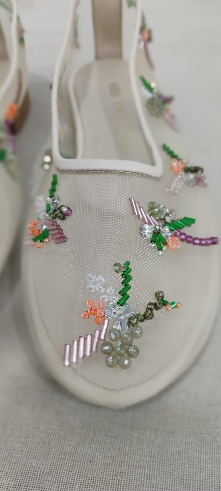 A close-up of a beige slip-on shoe decorated with colorful floral beadwork and sequins on a textured fabric background.