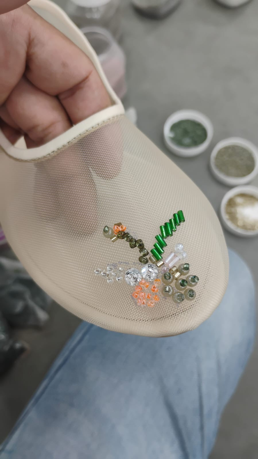A close-up of a hand holding a beige mesh shoe insert decorated with colorful rhinestones and a green spiral embellishment, with small containers of glitter or powder in the background.